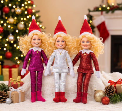 Three Dolly Parton - The Country Elf parody dolls in sparkly pink, silver, and red outfits displayed in a festive Christmas setting with a lit tree and holiday presents.